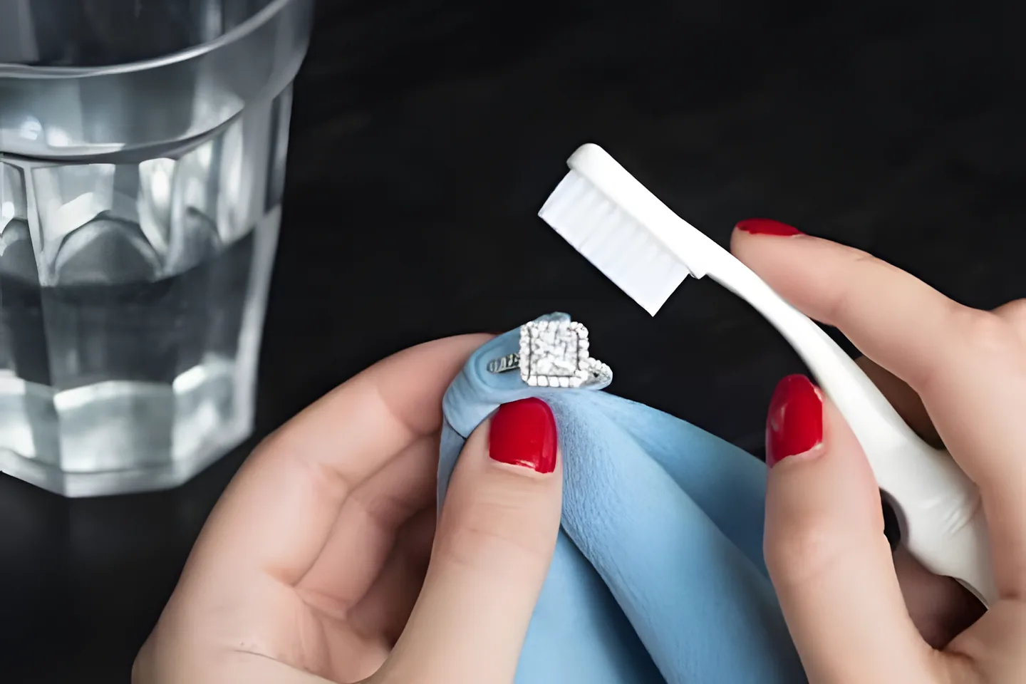 Hands cleaning a diamond ring with a soft cloth and a soft-bristled brush beside a glass of water.